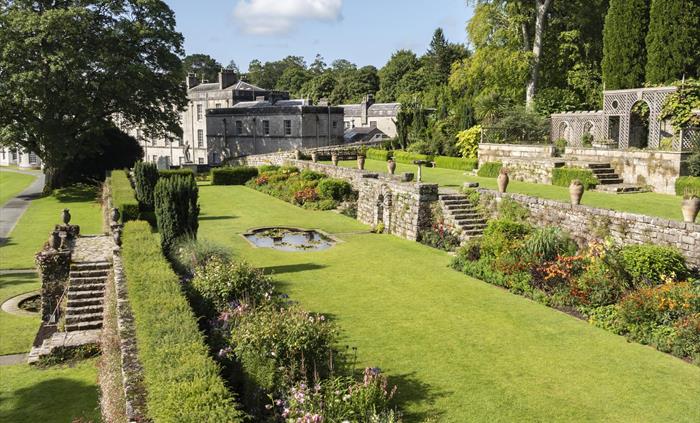 Terraced gardens with stone steps, colorful flower beds, and a small pond, set against a backdrop of tall trees and a large historic mansion under a s